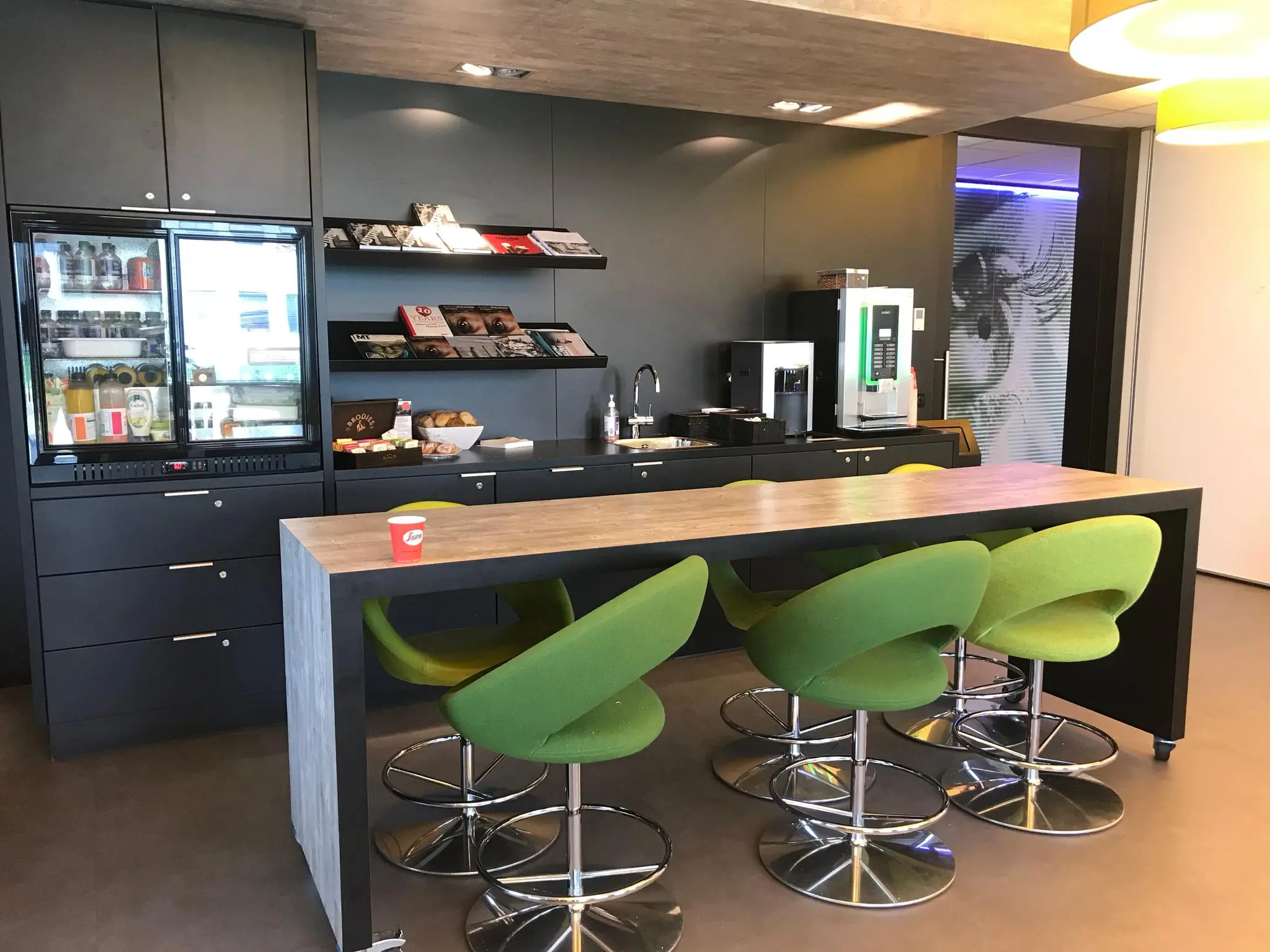 Moderne bedrijfskantine in Rivium Quadrant met groene barkrukken, een lange tafel, drankautomaat en tijdschriftenrekken.